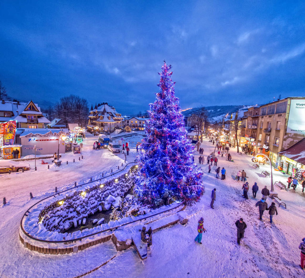 Veľký, osvetlený vianočný stromček na zasneženom námestí v Zakopanom počas vianočných trhov.