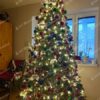 Vysoký umelý vianočný stromček, s tamvozelenými vetvičkami, husto ozdobený farbenými ozdobami a teplým osvetlením.