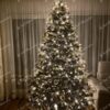 Zasnežený vysoký umelý vianočný stromček, husto ozdobený bielo-zlatými ozdobami s bielym osvetlením.