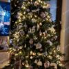 Sýtozelený hustý umelý vianočný stromček, ozdobený bielo-ružovými ozdobami a teplým bielym osvetlením, s bielym kobercom pod stromček