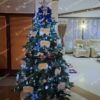 Umelý vianočný stromček so zelenými vetvičkami, ozdobený bielo-modrými ozdobami, s bielym kobercom pod stromček.