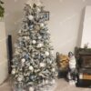 Husto zasnežený umelý vianočný stromček s modro-strieborno-bielými ozdobami a bielym koberčekom pod stromček.