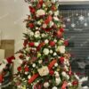 Umelý vianočný stromček so zelenými vetvičkami, husto ozdobený červeno-bielymi vianočnými ozdobami, zladený s bielym koberčekom pod stromček a vianočným Mikulášom