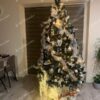 Umleý vianočný stromček so zasneženými koncami vetvičiek , ozdobený bielymi ozdobami a vianočnou stuhou s bielym koberčekom pod stromček.