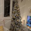 Zasnežený umelý úzky vianočný stromček so zlatými ozdobami a bielym koberčekom pod stromček.